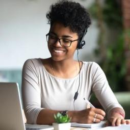 A person wearing headphones writes on paper while using a laptop.